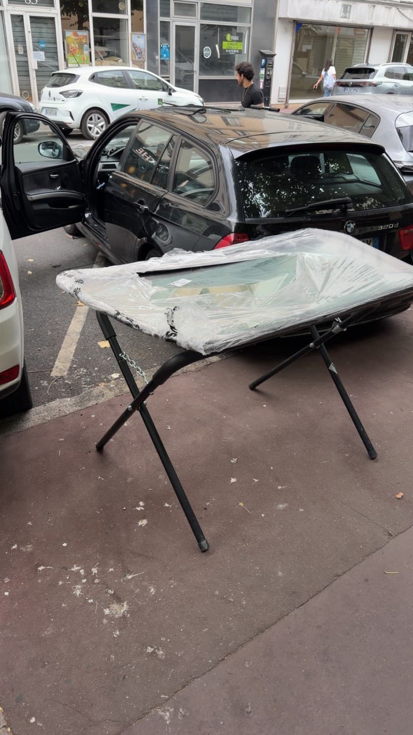 BMW Série 3 Touring noire, vitrage neuf déballé sur tréteaux, sur le trottoir à côté du véhicule